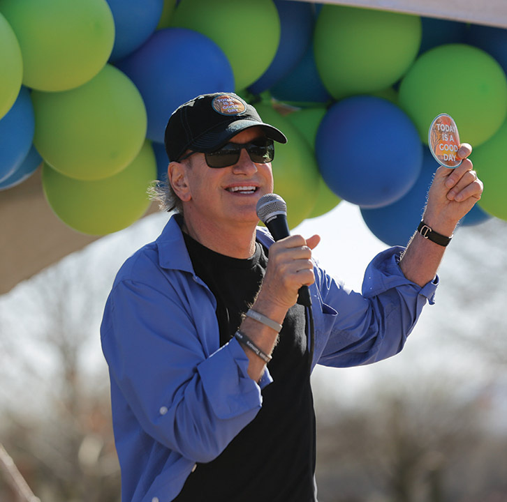 A white man with a black shirt under a long-sleeve blue shirt waves his hand while holding a microphone in front of green and blue balloons at an event. 