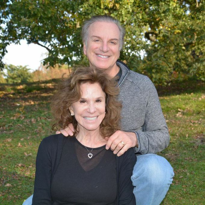 A white couple sits on a lawn with the woman, wearing a black sweater, leaning against her husband in a gray sweater and jeans.