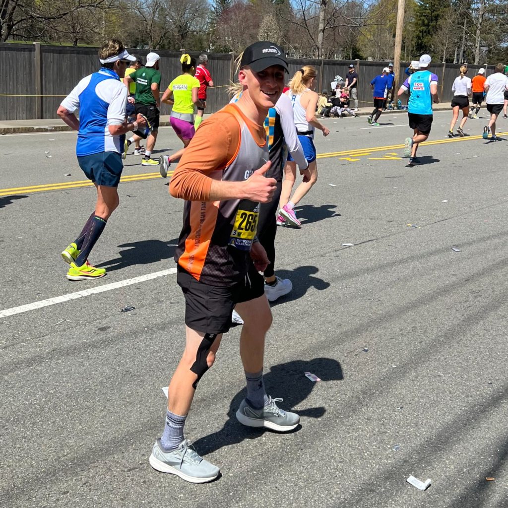 A 28-year-old man gives a thumbs up while on the Boston Marathon course. He is wearing an orange race shirt, black shorts, and gym shoes.