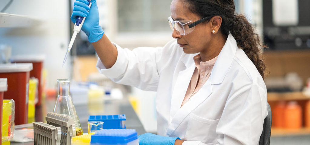 An ethnic female biochemist is working in a laboratory. She is seated at a work station and is using a pipette. The researcher is going to analyze the medical sample.