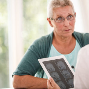 A female patient talks with a doctor as part of their informed decision-making. The doctor holds a tablet with brain imaging on it.