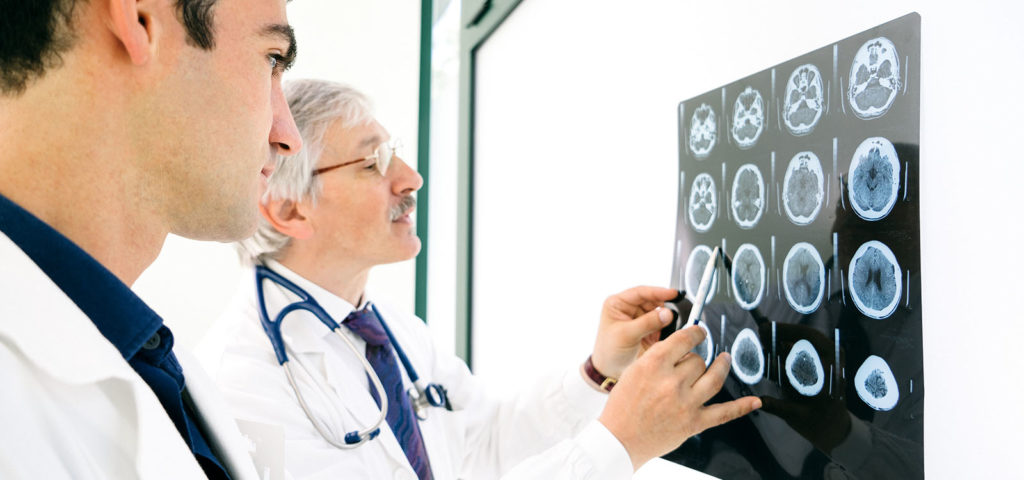 Two men in white coats examine medical imaging with the older man pointing at one scan with his pen.