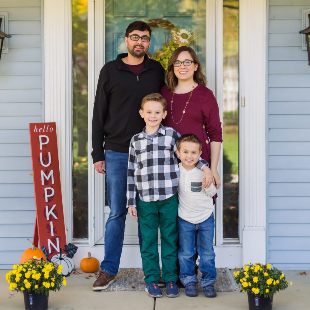 A family stands on their porch with mums and a "Hello Pumpkin" sign. Mom and dad are in their 30s and joined by two young boys.