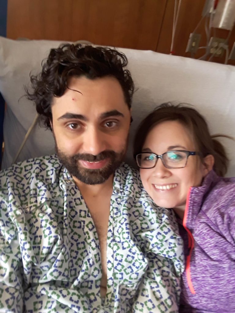 A bearded man sits in a hospital bed in a gown next to his wife in a purple leisure top and glasses.