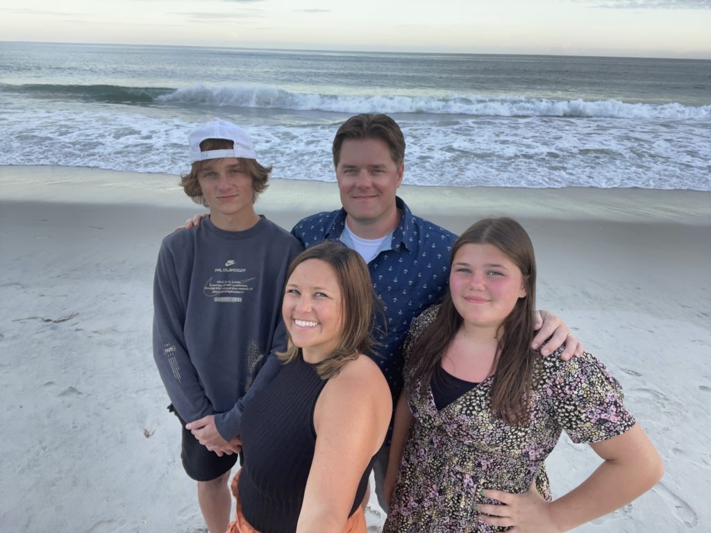 A four-person family stands on a beach at sunset, posing for a family selfie.