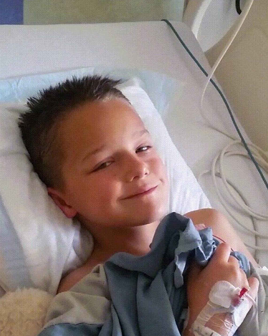 A young boy sits in a hospital bed wearing a hospital gown, hooked up to IVs, and smirking at the camera.