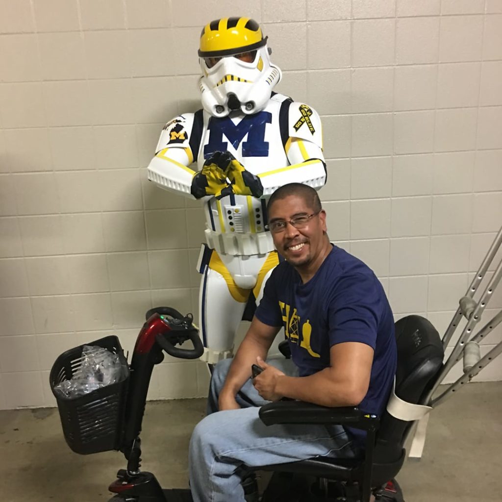 A man sits in his scooter, crutches hanging off the back, in front of a University of Michigan stormtrooper.