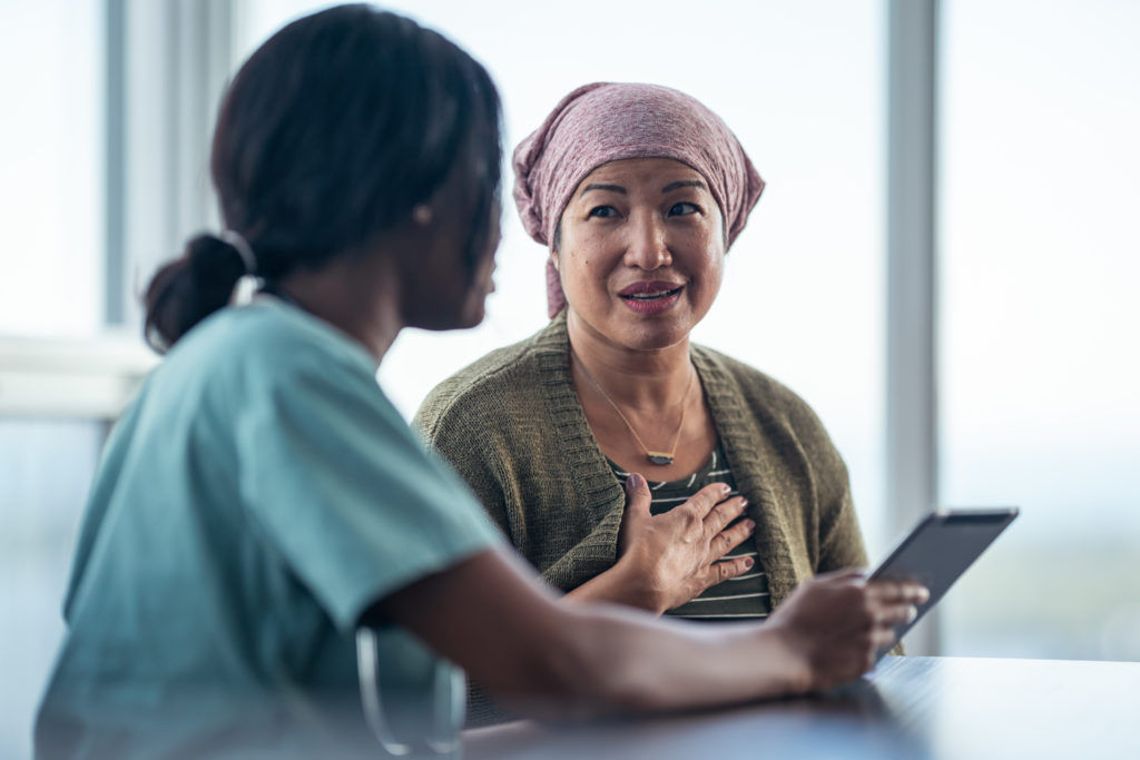 An Asian woman with cancer is consulting her doctor. The two women are seated at a table together. The patient is wearing a bandana to hide her hair loss. The medical professional is showing the patient test results on a digital tablet. They are discussing a treatment plan.