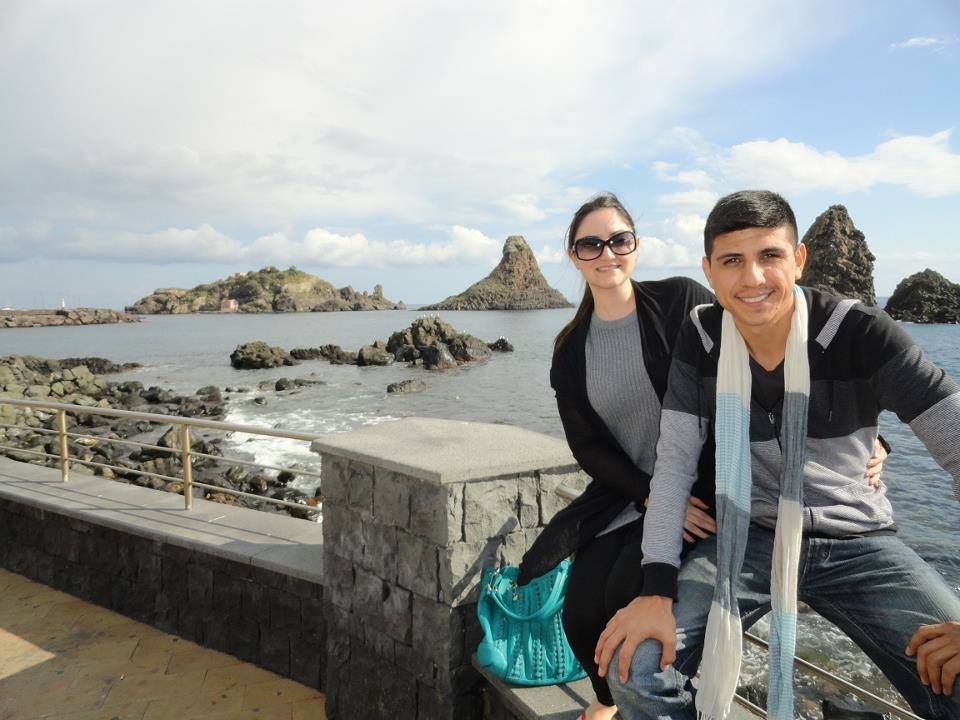 A man in a monochrome sweater, jeans, and scarf sits on a railing alongside a women in a gray shirt and black cardigan on vacation.