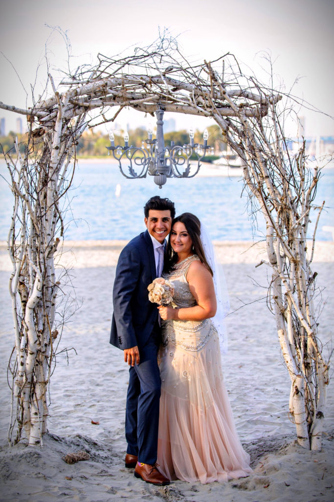 A man in a blue tuxedo and a woman in an ivory dress stand under a driftwood arch with a chandelier after getting married on the beach.