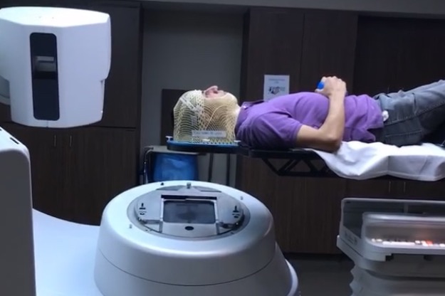 A man in a purple shirt and jeans wears a radiation mask and lays down while receiving radiation treatment for a brain tumor.