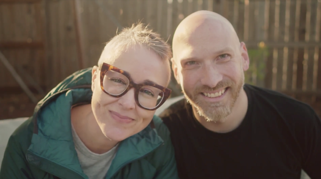 A woman with short hair following a craniotomy and glasses smiles next to a smiling man with a shaved head and beard..