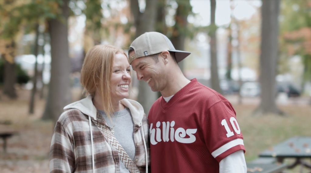 A woman with red hair wears a plaid jacket and stands next to a man in a Phillies jersey and a backwards hat.