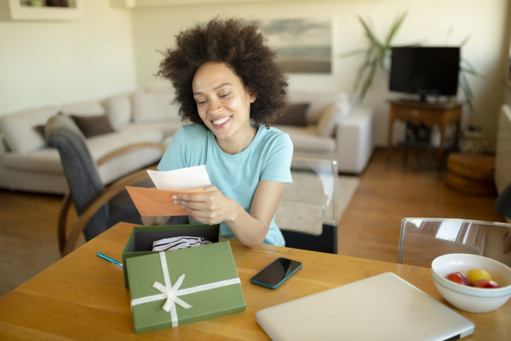 Cheerful African American woman with a beautiful smile, opening a gift at home, feeling happy and reading a letter she received with it