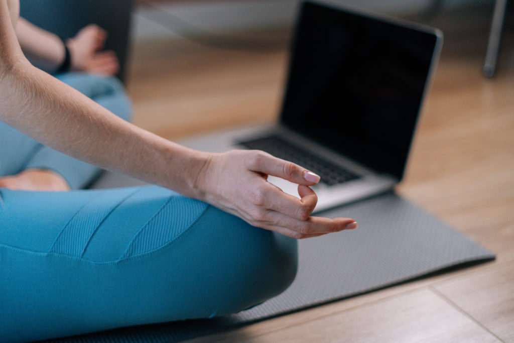 Close-up of unrecognizable young woman meditating at home sitting in lotus position on yoga mat near laptop computer holding hands on knee in Om position.