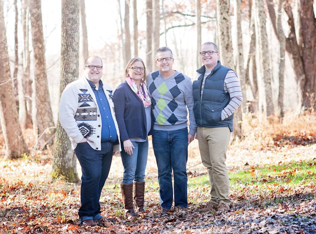 Two adult sons stand on either side of their parents — all wearing sweaters — for a family portrait outdoors with leafless trees in the background.
