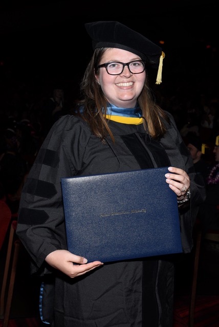 A woman with glasses smiles for a photo in a cap & gown, holding up her diploma.