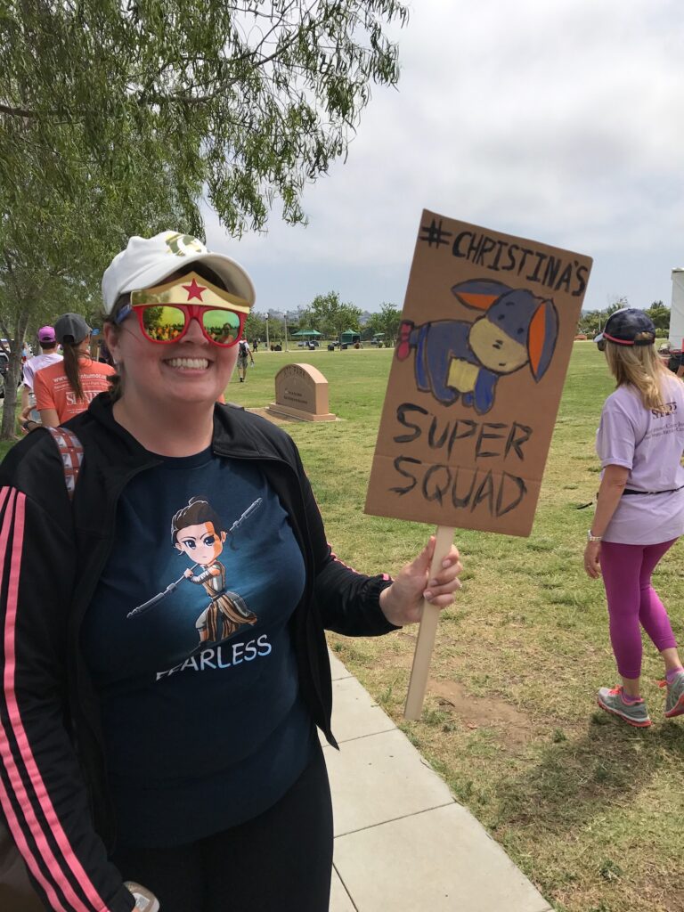 A woman in sunglasses and a hat walks holding up a #Christin'a Super Squad sign.