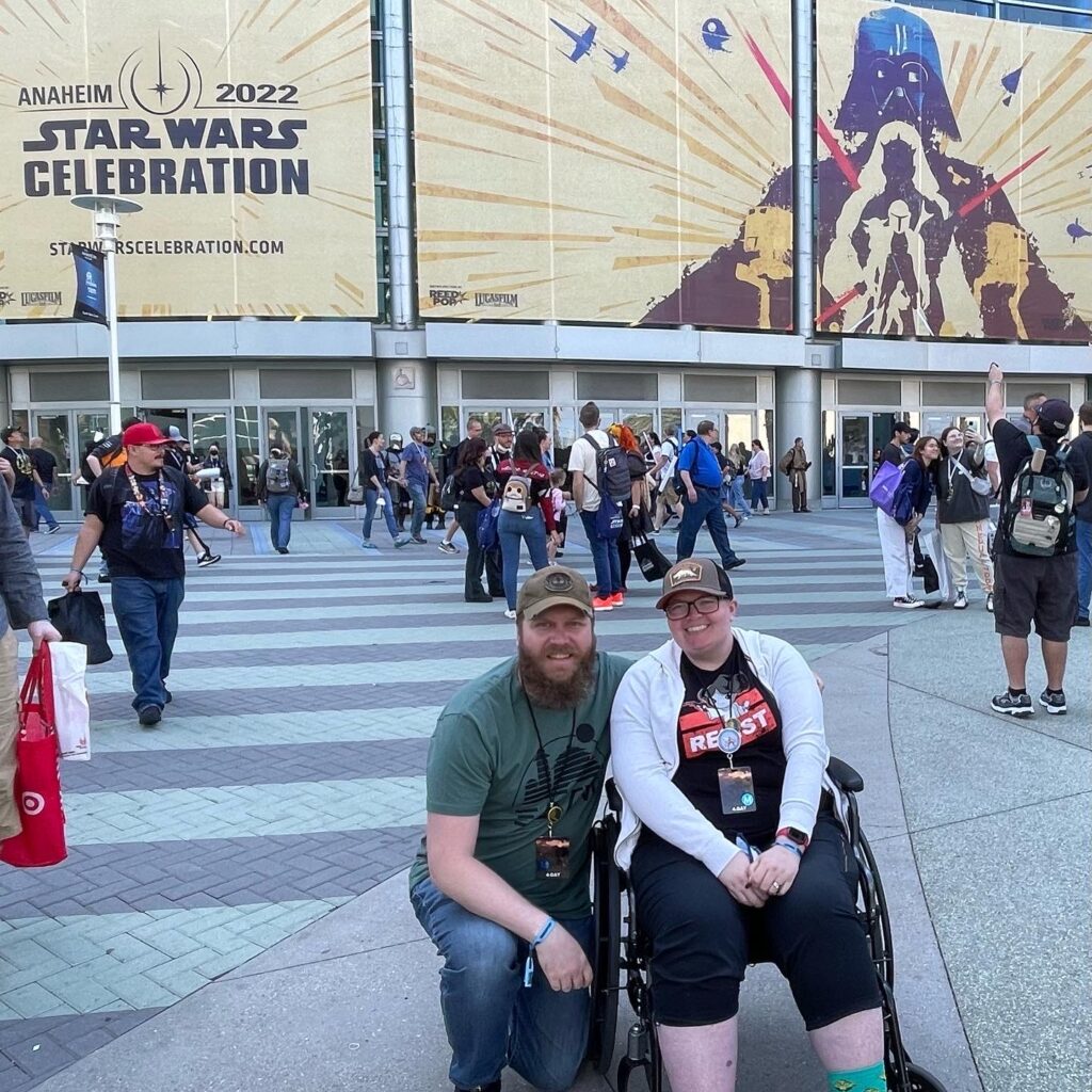 Woman sits in wheelchair with husband kneeling beside her in front of Star Wars Celebration marquee.