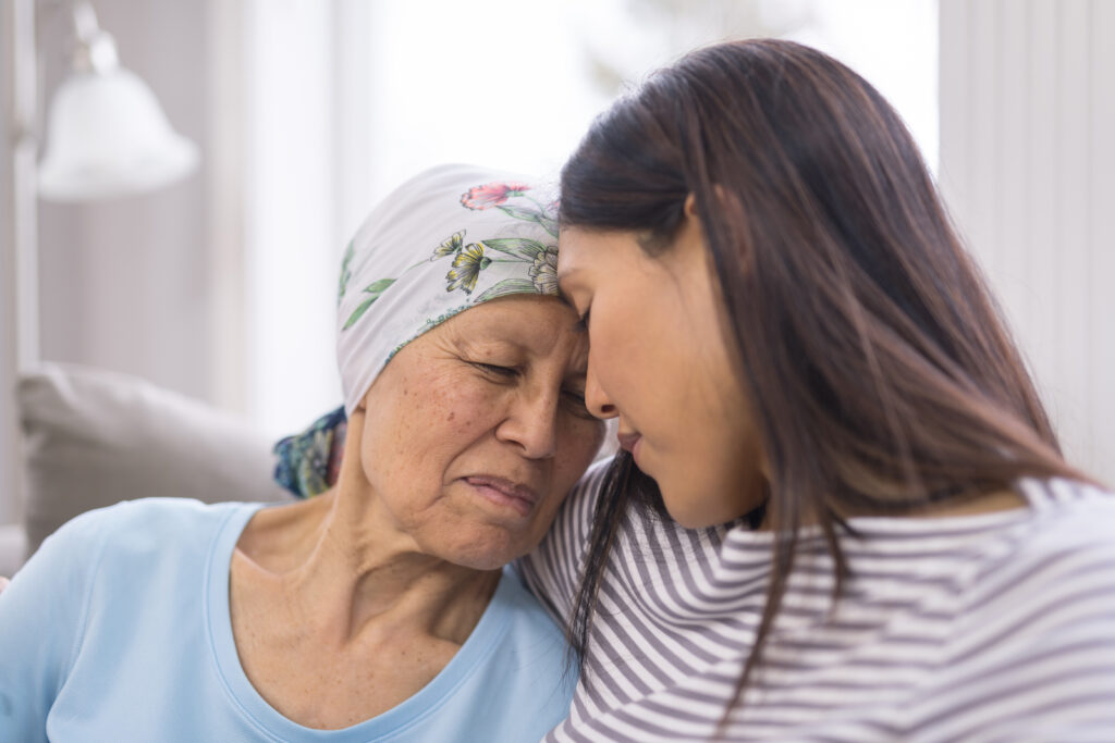 Asian elderly woman with cancer and wearing a headcovering is embracing her adult daughter. They are sitting on a couch and their foreheads are touching.
