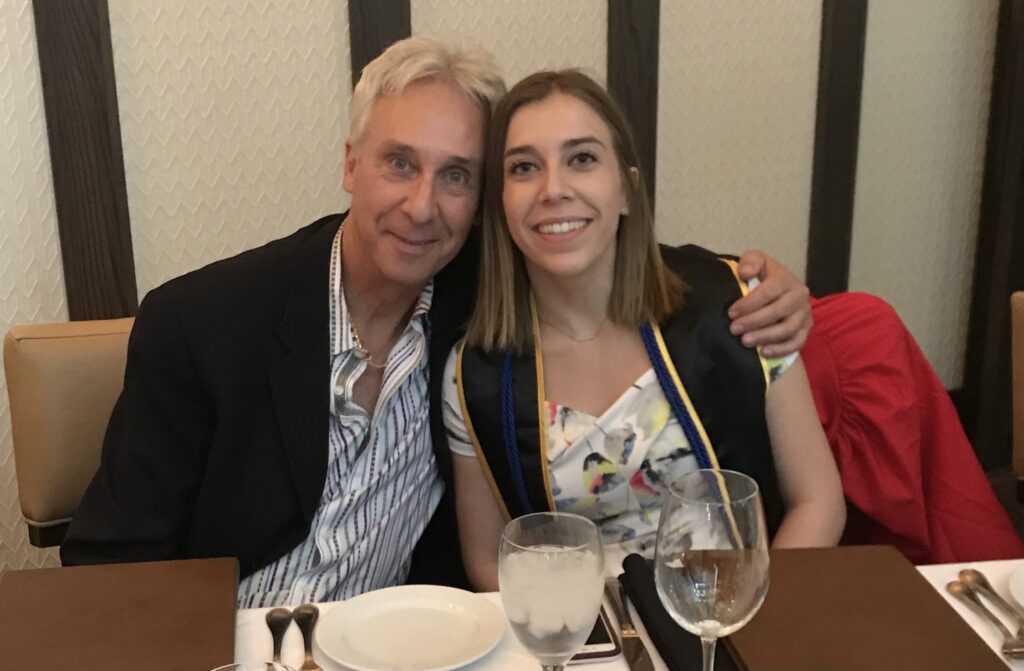 A young woman sits next to her dad in his 60s with his arm around her at a restaurant.
