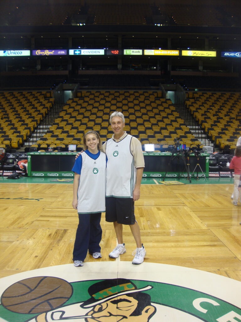A young adult woman stands next to her father on the Boston Celtics court.