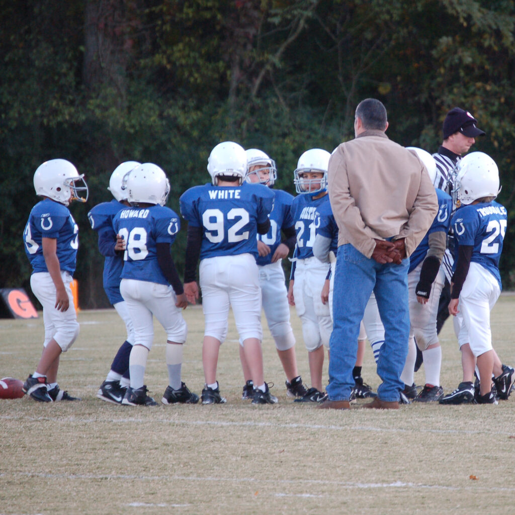 A pee wee football team gathers on the field with their coach looking over.