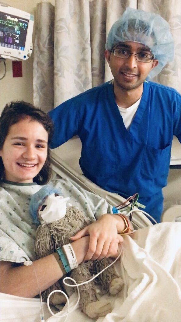 A college student is in a hospital bed, posing for a photo with a health care professional in blue scrubs and hair net, before her brain surgery.