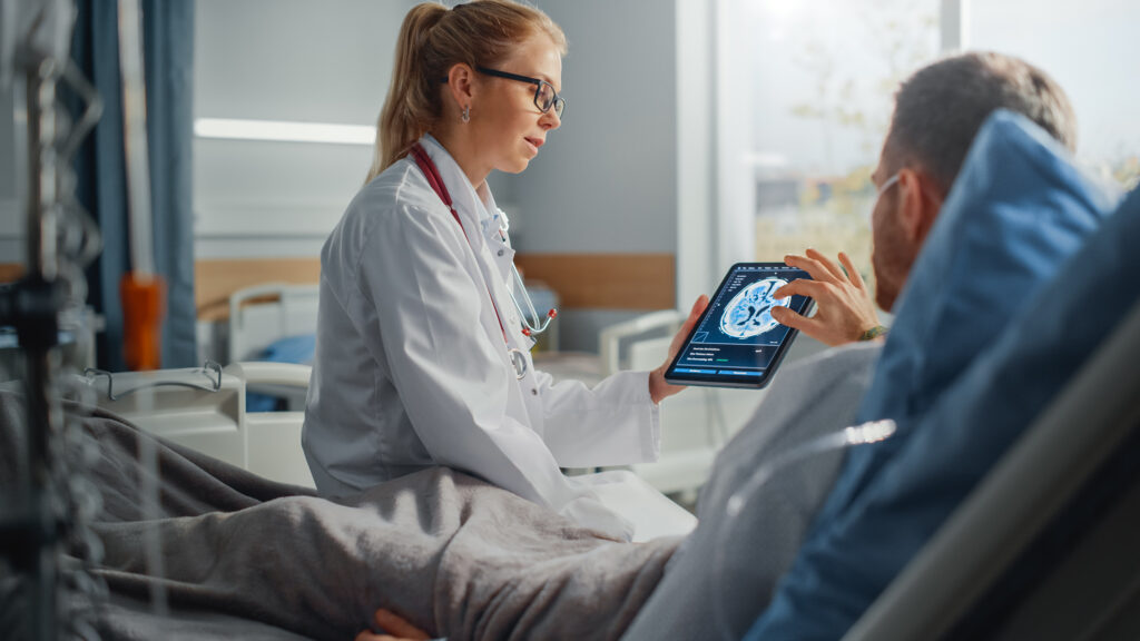 Hospital Ward: Friendly Female Doctor Talks to Male Patient Resting in Bed, Uses Tablet Computer, Shows and Explains Brain Scan Results During Brain Tumor Second Opinion