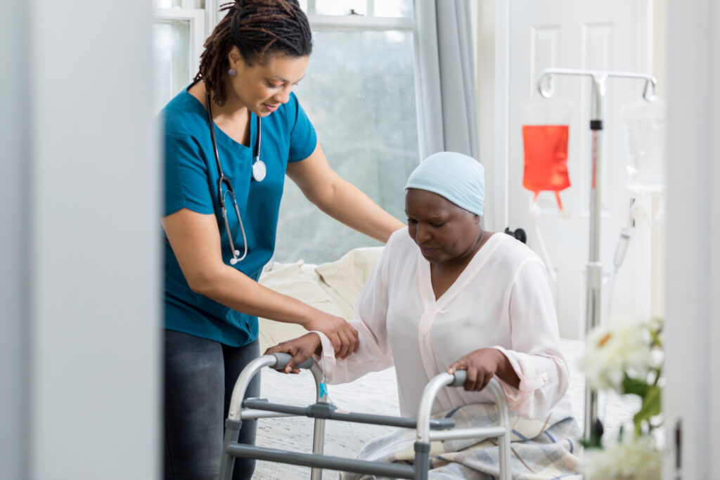 Young female home healthcare professional helps a patient get out of bed by using a walker after surgery. The woman is wearing a cap on her head. An iv drip is in the background.