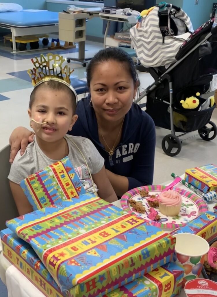 A young girl, who is also a pediatric brain tumor patient, sits next to her mother with presents and birthday cake in front of them.