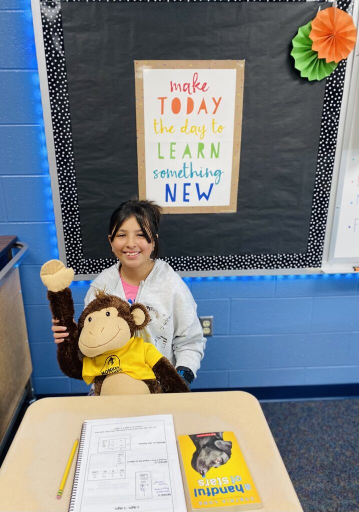 An elementary student, who is also a pediatric brain tumor patient, sits in her classroom holding a stuffed monkey.