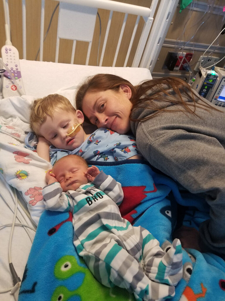 A young brain tumor patient rests in his hospital bed alongside mom and newborn baby brother.