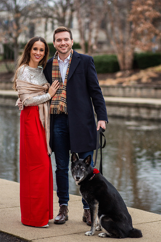 A couple smiles by a pond with their dog for a holiday family photo.