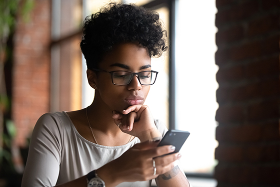 Serious black woman wearing glasses holding phone browse internet, which can contribute to cognitive fatigue.
