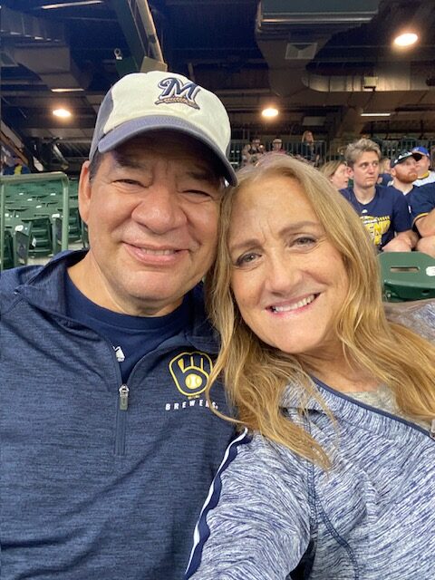 A couple smiles at a Milwaukee Brewers game. Jeanne discusses caregiving and loss after her husband's glioblastoma diagnosis.