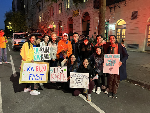 Twelve people stand, some with signs, to cheer on a woman running in the NYC Marathon in honor of her father living with a brain tumor.