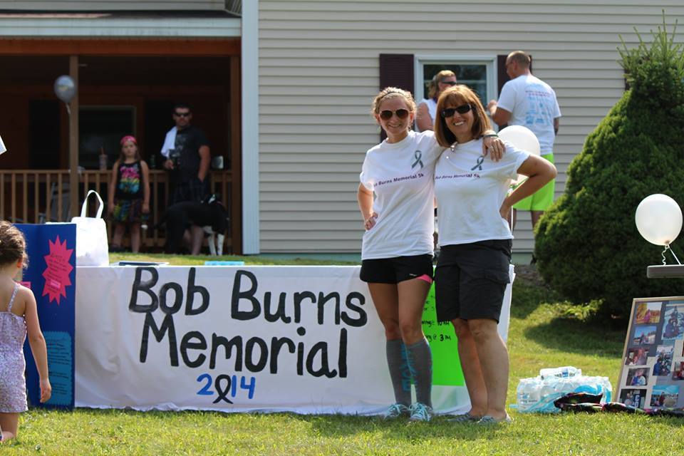 Two women in white t-shirts with gray ribbons stand in front of a Bob Burns Memorial 5K 2014 banner.