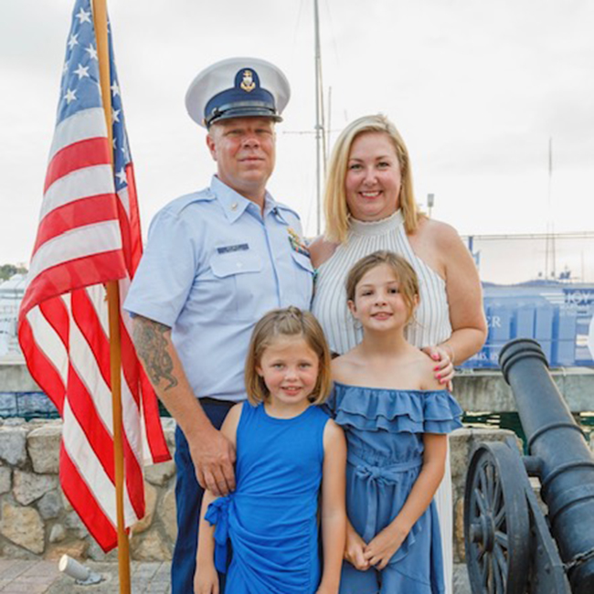 A male Coast Guard service member stands with his wife and two young daughters next to an American flag and canon. 