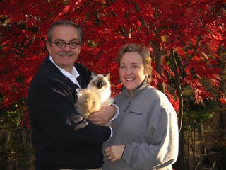 A man in glasses holds a cat while standing alongside his wife in front of a tree with bright red leaves.