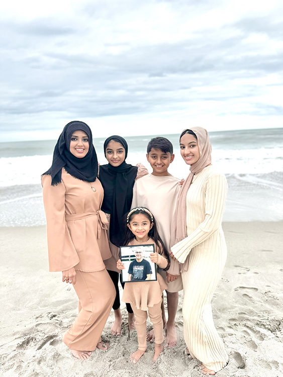 A little girl holds up a photo of her father, who passed away after a glioblastoma diagnosis, alongside her family standing on a beach.