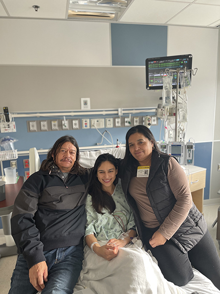A female patient with central neurocytoma smiles in a hospital bed alongside her two parents, one on each side.
