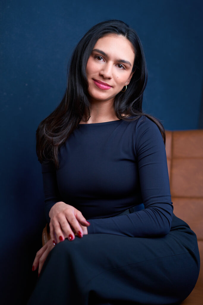 A woman in a dark blue dress sits on a chair for a professional headshot.