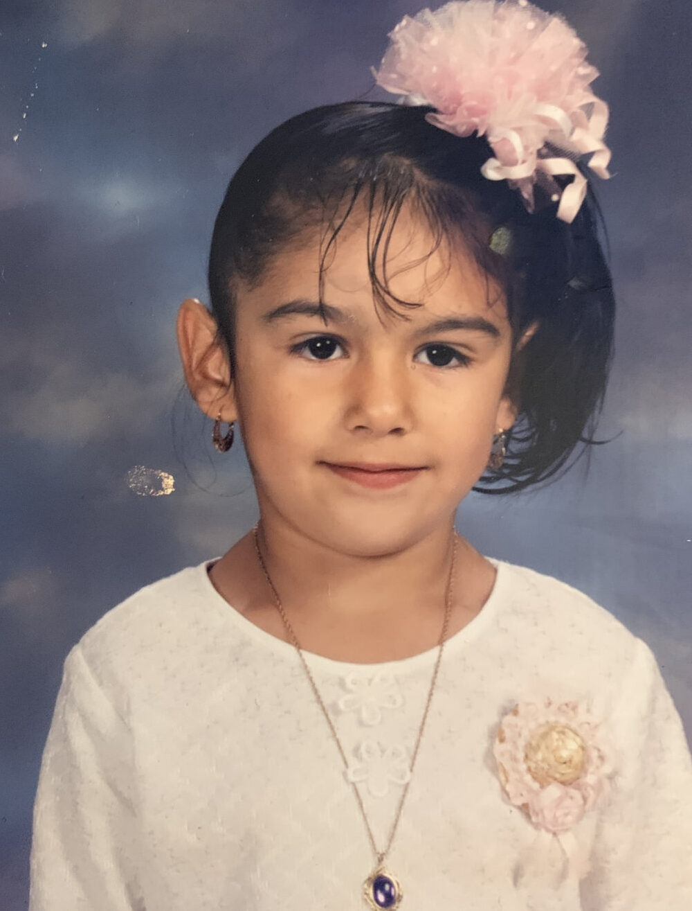 A young girl with a side ponytail and a pink bow up top smiles for a school photo.