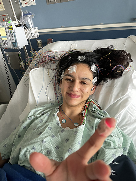 A woman with central neurocytoma smiles in a hospital bed and gown with nodes on her head and hand raised toward camera.