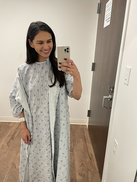 A smiling woman, standing in a hospital gown, holds her phone up to take a photo in a mirror.