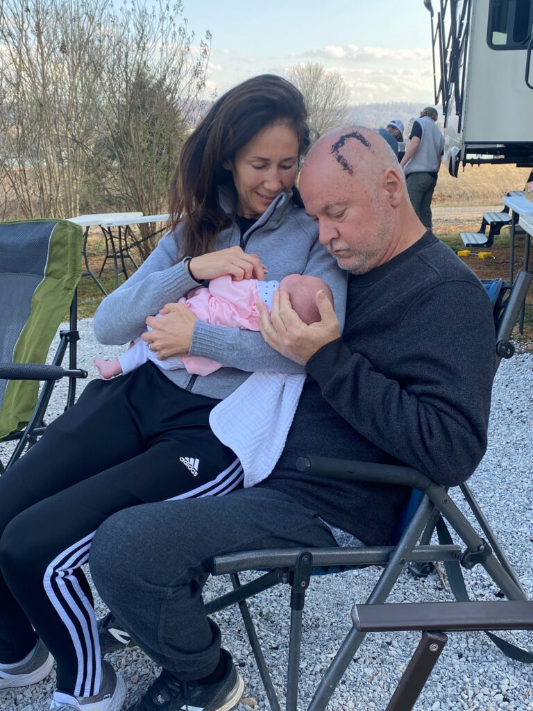 A man with a fresh craniotomy scar and woman hold their infant grandchild.