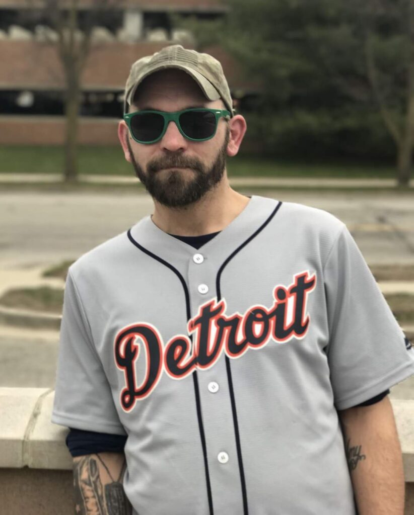 A bearded man stands wearing a Detroit baseball jersey, baseball hat, and sunglasses.