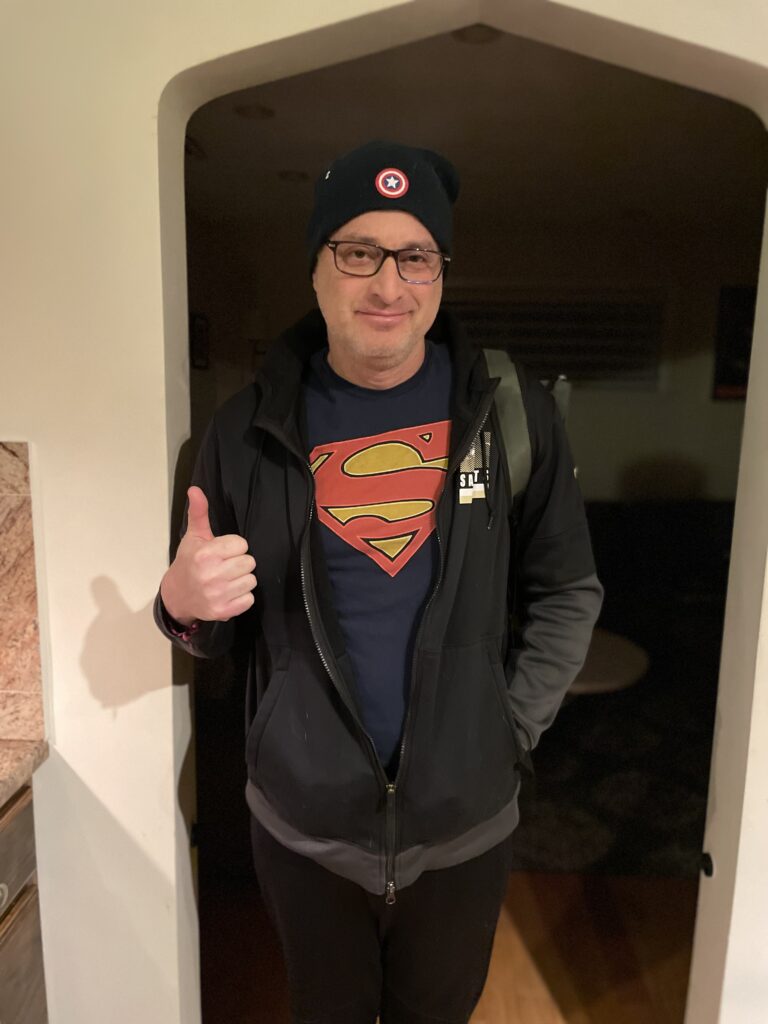 A man wearing a Superman t-shirt, light jacket, and a Marvel winter knit hat gives a thumbs up to the camera