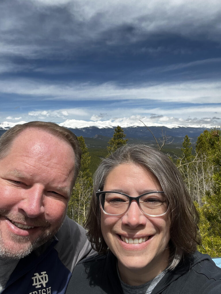 A couple stand on mountainous terrain with a forest behind them and mountains in the distance.